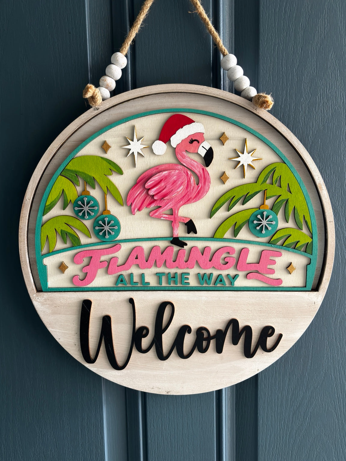 Pink flamingo in a Santa hat between palm trees with ornaments and Flamingle All the Way lettering, displayed in welcome sign base