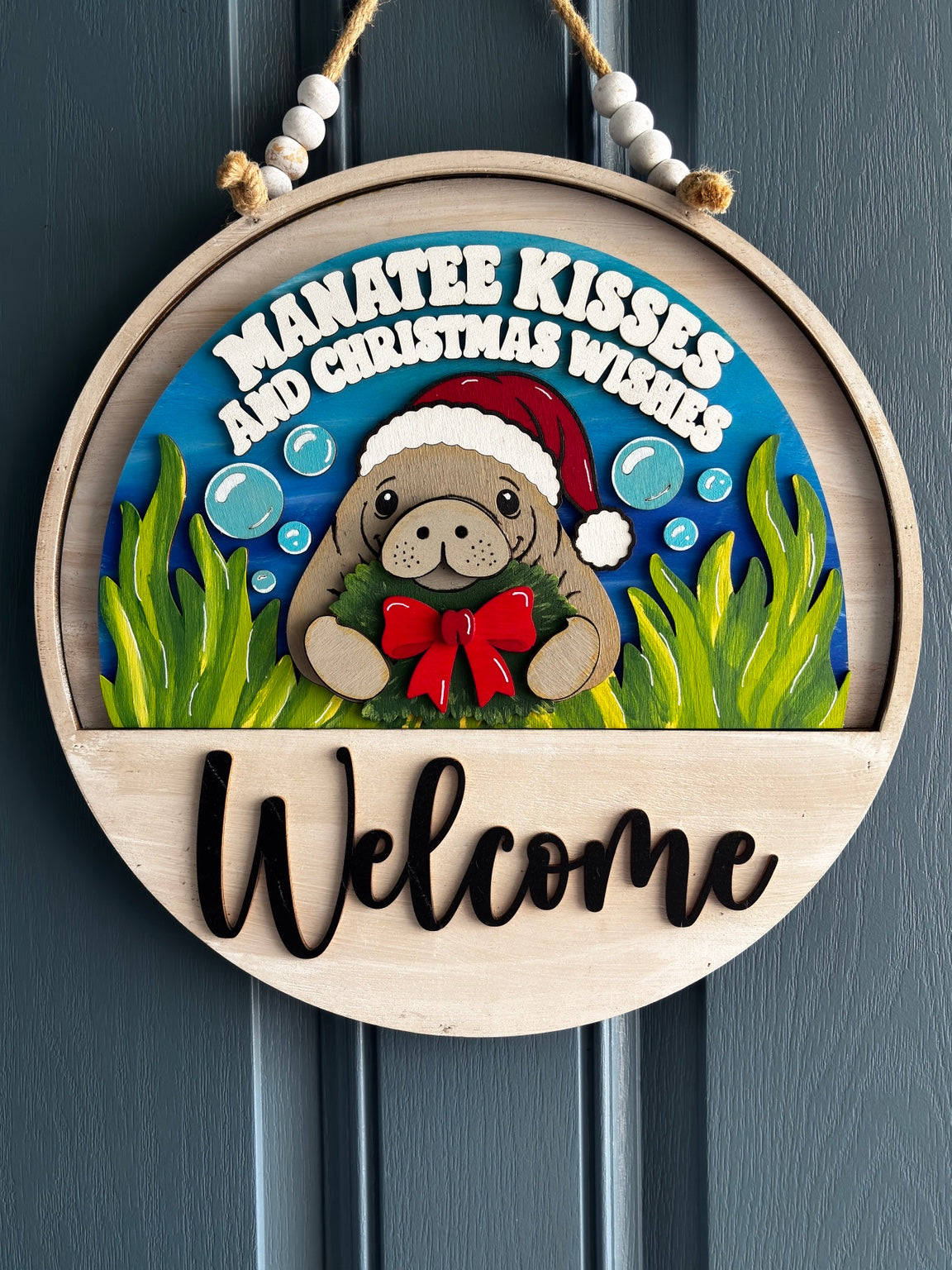 Manatee in a Santa hat holding a Christmas wreath, with bubbles, seagrass and Manatee Kisses and Christmas Wishes lettering, displayed in welcome sign base